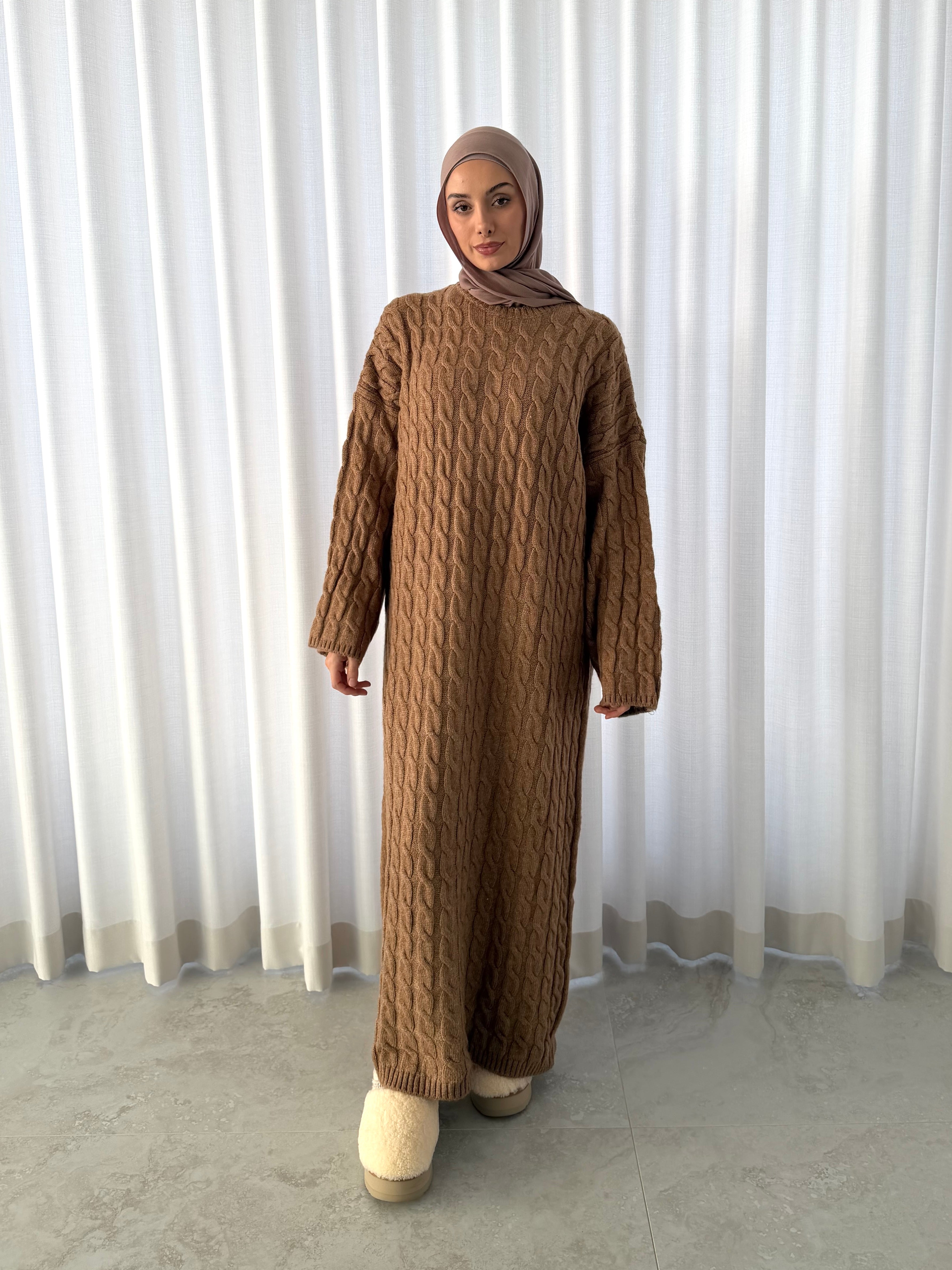 Model wearing a modest a camel coloured cable wool dress with long sleeves and a relaxed oversized fit. Styled with cream fluffy slippers against a neutral curtain backdrop.