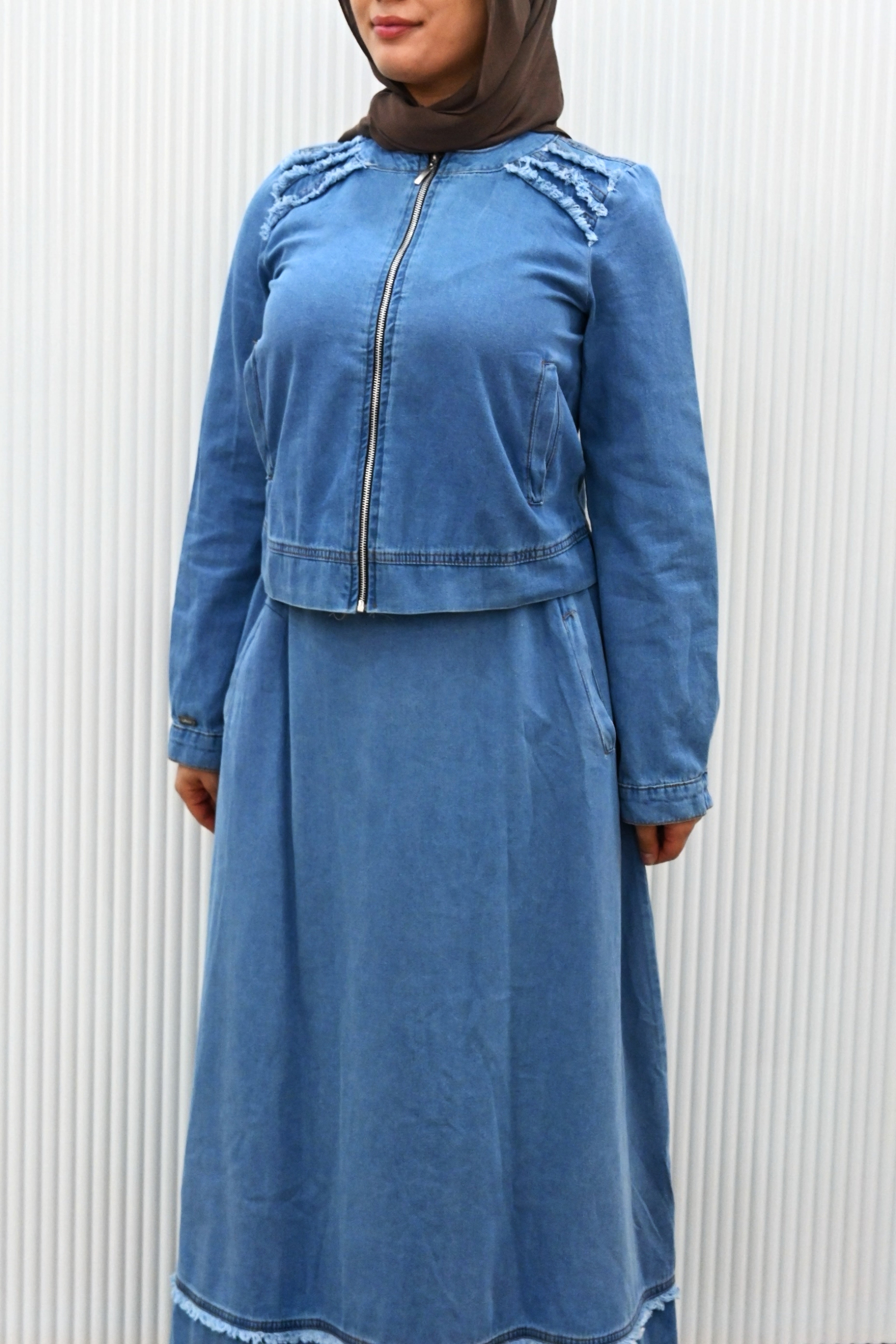 This is a two-piece modest denim-style outfit featuring a long A-line skirt and a matching zip-up jacket. The skirt has layered frayed hem details for a casual chic look, while the jacket is designed with full sleeves and light embroidery accents on the shoulder. Paired with a plain inner top, this set combines comfort and style, making it perfect for everyday wear or casual outings.