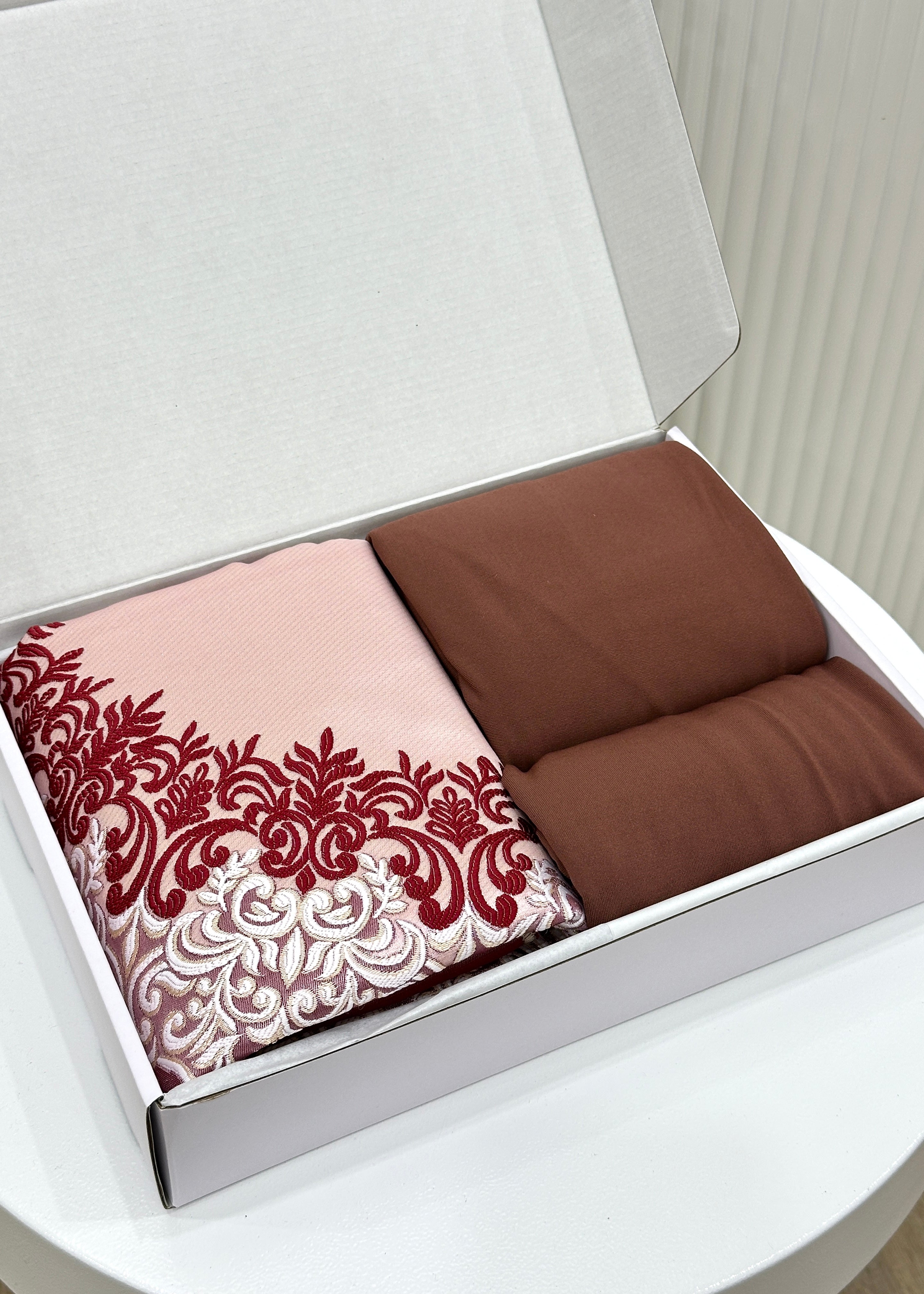 A gift box open to reveal a prayer mat and a prayer set with a dusty rose color scheme, including a decorative pattern on the prayer mat.