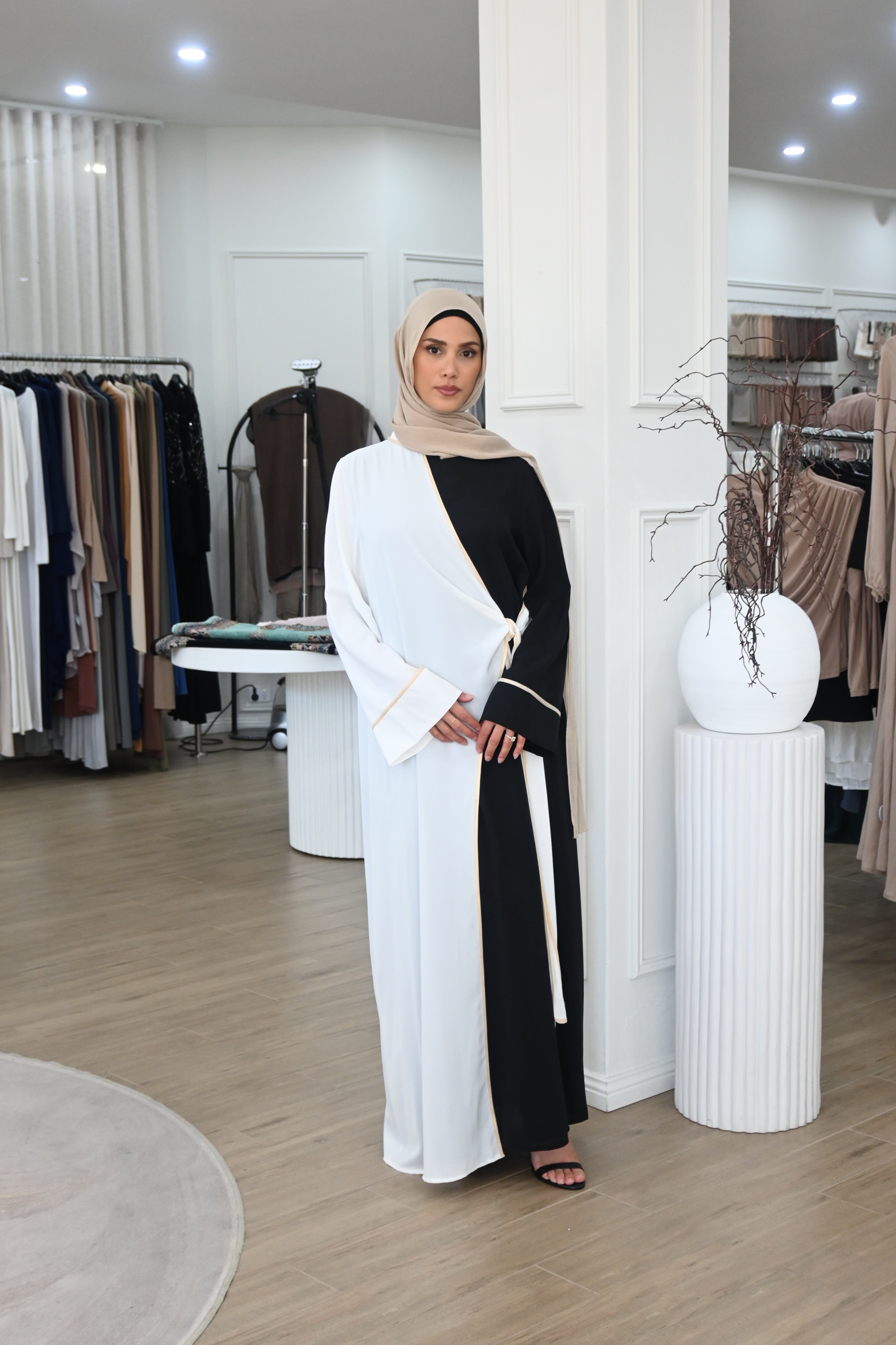 A modern reinterpretation of classic modest wear. This dual-tone abaya features a black base with a soft ivory overlay, delicately trimmed with subtle gold piping. The wrap design ties gracefully at the side, creating an effortless layered effect. Its fluid drape and timeless contrast make it ideal for both daywear sophistication and evening elegance.