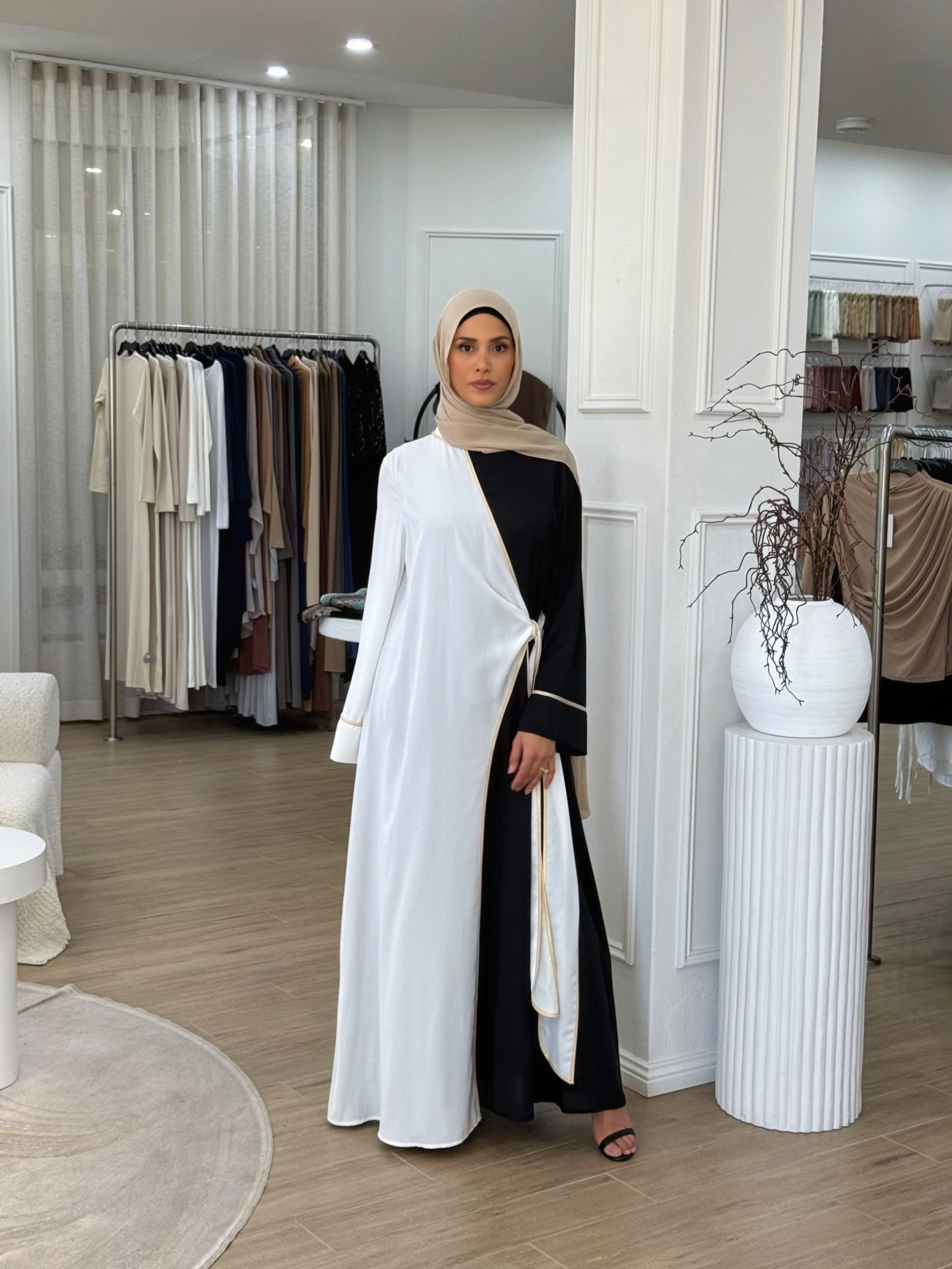 A modern reinterpretation of classic modest wear. This dual-tone abaya features a black base with a soft ivory overlay, delicately trimmed with subtle gold piping. The wrap design ties gracefully at the side, creating an effortless layered effect. Its fluid drape and timeless contrast make it ideal for both daywear sophistication and evening elegance.