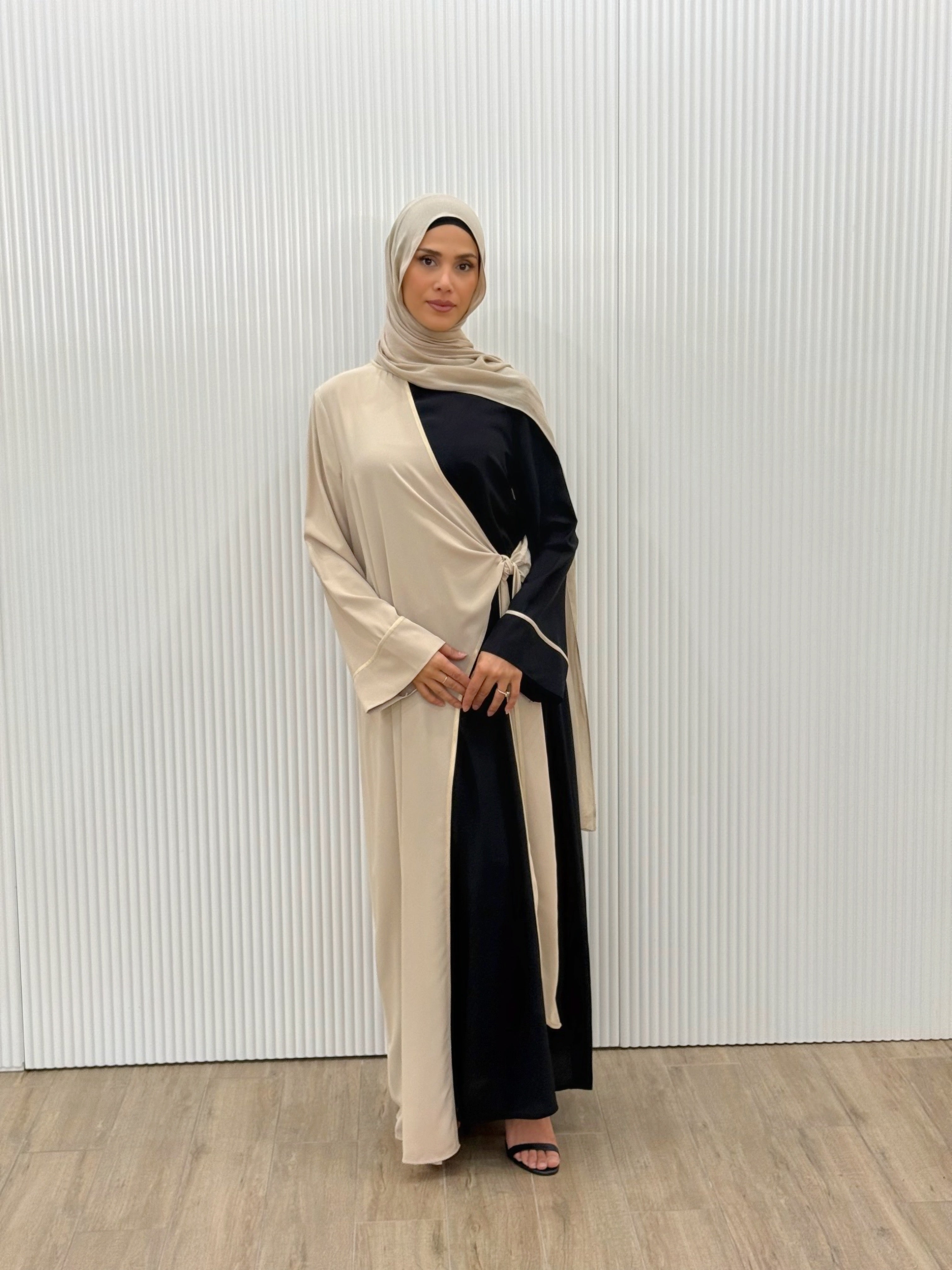 A modern reinterpretation of classic modest wear. This dual-tone abaya features a black base with a soft ivory overlay, delicately trimmed with subtle gold piping. The wrap design ties gracefully at the side, creating an effortless layered effect. Its fluid drape and timeless contrast make it ideal for both daywear sophistication and evening elegance.