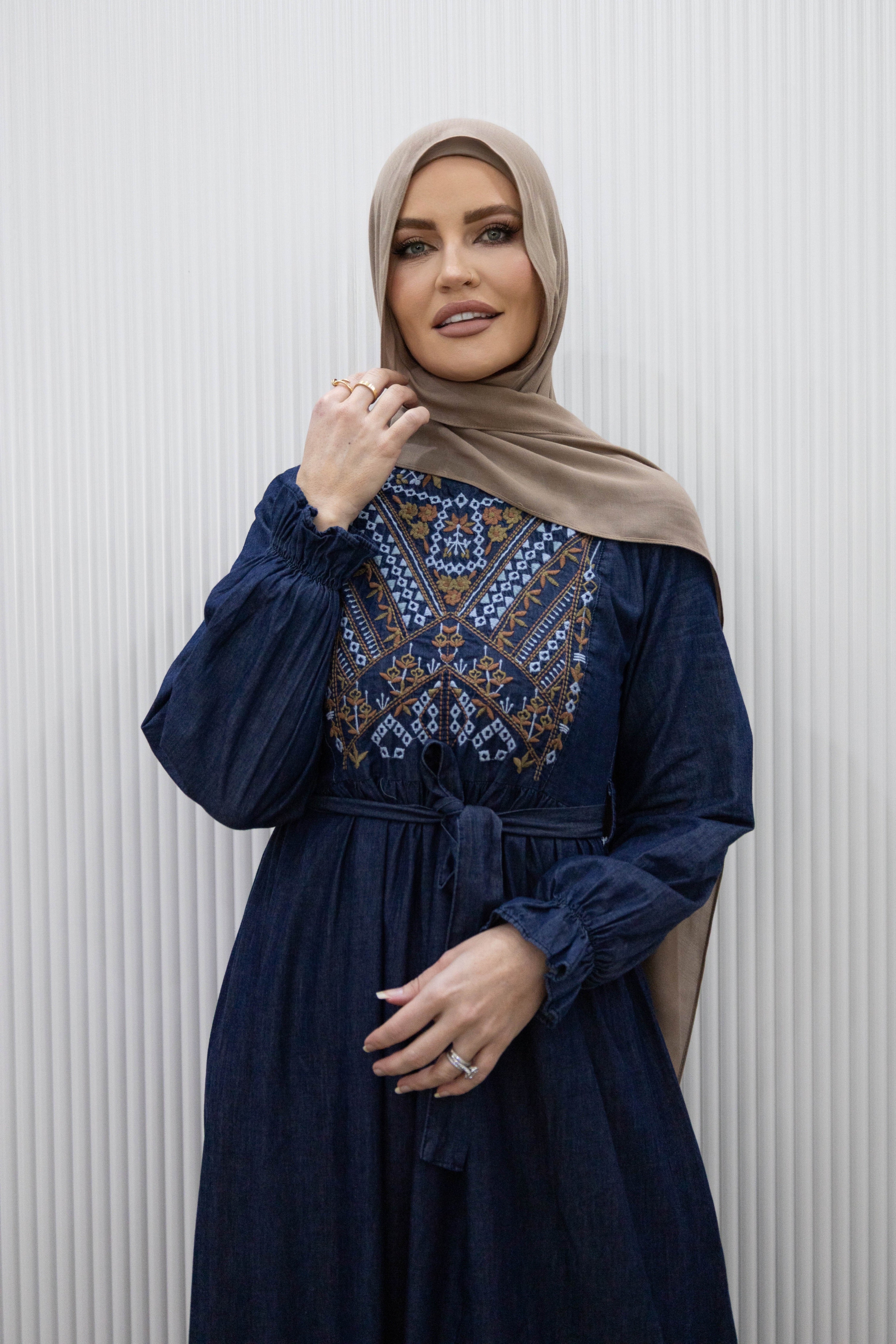 Taseem Dress in lightweight denim-style fabric with embroidered paisley design, long frill-cuffed sleeves, waist tie, and flared maxi skirt for a flattering, elegant silhouette.