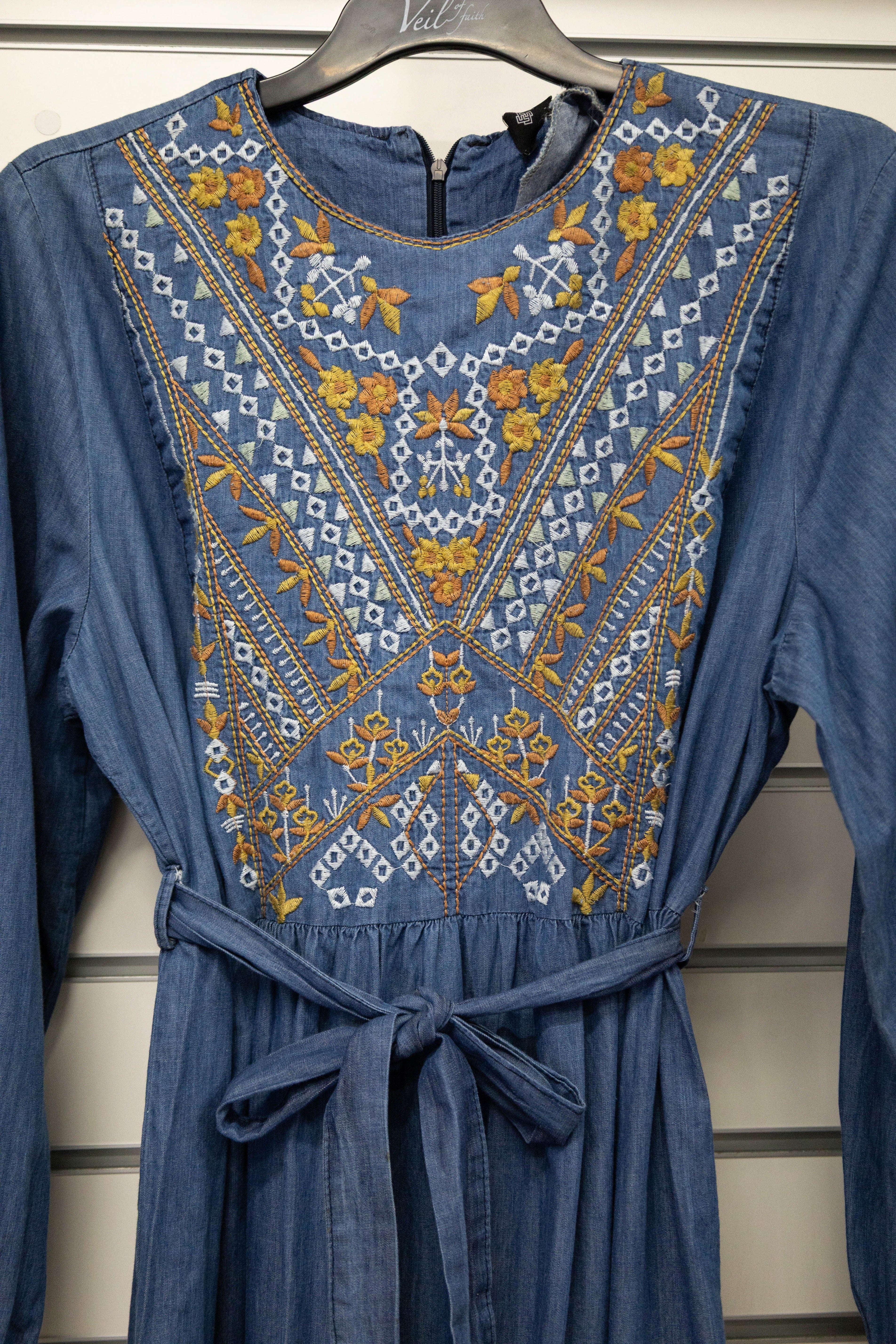 Taseem Dress in lightweight denim-style fabric with embroidered paisley design, long frill-cuffed sleeves, waist tie, and flared maxi skirt for a flattering, elegant silhouette.